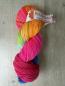 Preview: Ein handgefärbter Strang Sockenwolle von Herzfasern in kräftigen Neonfarben. Die Färbung kombiniert leuchtendes Pink, Orange, Gelb, Grün und Blau mit dunklen Sprenkeln. Der Strang liegt auf hellem Holzuntergrund und trägt eine Herzfasern-Banderole.