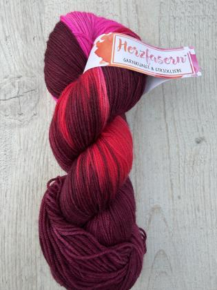 Ein handgefärbter Garnstrang in dunklen Beerentönen und Rot mit leuchtenden Pinkakzenten liegt auf einem hellen Holzuntergrund. Die Farben wirken tief und harmonisch mit lebendigen Highlights.