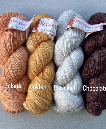 Vier Stränge handgefärbte Wolle liegen nebeneinander. Von links nach rechts: ein warmes Apricot-Orange (Solana), ein strahlendes Goldgelb (Golden), ein helles cremefarbenes Weiß (Perlmutt) und ein tiefes, schokoladenbraunes Garn (Chocolate).