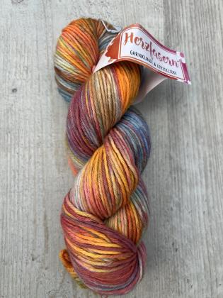 Ein handgefärbter Strang Wolle in warmen Herbstfarben – Orange, Rot, Gold und dezenten Blautönen. Das Garn liegt locker gedreht auf hellem Holz, das Etikett zeigt das Herzfasern-Logo.
