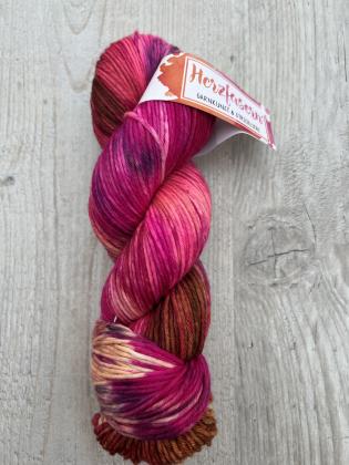 Ein Strang handgefärbter Wolle in kräftigen Pink-, Beerenton- und Kupfernuancen liegt auf hellem Holz. Der Strang ist gedreht, sodass die Farbverläufe sichtbar sind. Die Banderole von „Herzfasern“ ist umgelegt.