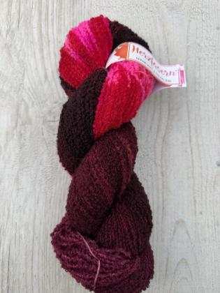 Ein handgefärbter Bouclé-Garnstrang in dunklen Beerentönen, Rot und leuchtendem Pink liegt neben einem gewickelten Knäuel auf hellem Untergrund. Die typische schlaufenartige Struktur des Garns ist deutlich sichtbar.