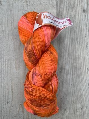Ein handgefärbter Strang Wolle in kräftigen Orange-, Kupfer- und Pinktönen mit feinen dunklen Sprenkeln. Der Strang liegt auf heller Holzoberfläche, das Etikett zeigt das Herzfasern-Logo in Pink und Orange.