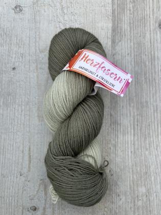 Ein handgefärbter Strang Wolle in gedeckten Grautönen – von hellem Steingrau bis zu dunklerem Taupe verlaufend. Der Strang liegt auf hellem Holz, das Etikett trägt das Herzfasern-Logo in Pink-Orange.