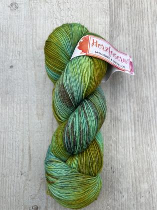Ein handgefärbter Strang Wolle in Moos- und Grüntönen mit feinen Braunsprenkelungen. Das Garn liegt auf heller Holzoberfläche, das Etikett zeigt das Herzfasern-Logo in Pink-Orange.