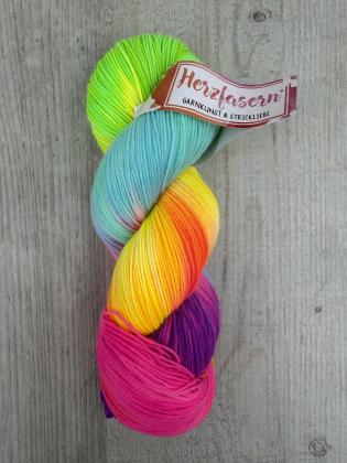 Handgefärbter Wollstrang in kräftigen, leuchtenden Farben. Der Strang zeigt einen Verlauf aus Neon-Grün, Gelb, Orange, Türkis, Pink und Violett. Die Wolle liegt auf einem hellen Holzuntergrund und trägt eine Herzfasern-Banderole.