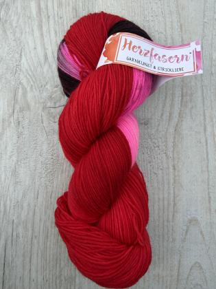 Ein handgefärbter Garnstrang in kräftigem Rot mit leuchtenden Pinkanteilen und dunklen Akzenten liegt auf einem hellen Holzuntergrund. Die Farben wirken intensiv und kontrastreich.