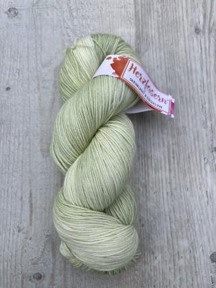 Ein handgefärbter Strang Wolle in zartem, gleichmäßigem Pistaziengrün. Der Strang liegt auf heller Holzoberfläche, das Etikett zeigt das Herzfasern-Logo in Pink und Orange.