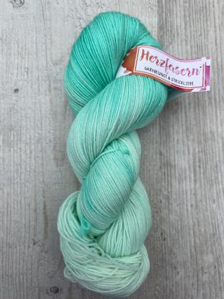 Handgefärbter Strang in mintgrünen und eisig hellen Tönen, weich verlaufend, fotografiert auf hellem Holzuntergrund mit Herzfasern-Etikett.