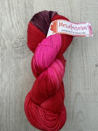 Ein handgefärbter Garnstrang in kräftigem Rot mit Übergängen zu leuchtendem Pink und Rosétönen liegt auf einem hellen Holzuntergrund. Die Farbverläufe sind weich und lebendig.