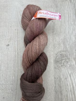 Ein Strang handgefärbter Wolle in warmen Schoko- und Rosétönen liegt auf hellem Holz. Der Strang ist gedreht, die natürliche Melierung und die weichen Farbverläufe sind deutlich sichtbar. Die Herzfasern-Banderole ist teilweise zu sehen.