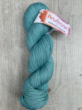 Ein Strang handgefärbtes Garn in der Farbe Sage. Das Garn zeigt ein klares, kühles Grün mit feinem Schimmer. Die Banderole trägt das Herzfasern-Logo. Der Strang liegt auf hellem Holzuntergrund.