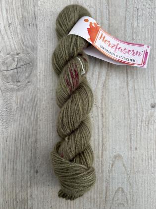 Ein handgefärbter Strang Herzfasern No. 94 – Cashmere Cloud in der Farbe Olive gesprenkelt. Zartes Olivgrün mit feinen Sprenkeln in Pink, Gold und Braun. Fotografiert auf hellem Holz mit Herzfasern-Banderole.