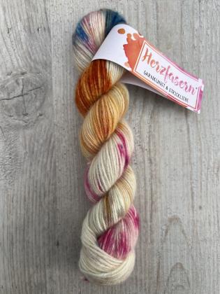 Ein handgefärbter Strang Herzfasern No. 94 – Cashmere Cloud in der Farbe Eleganz der Zeit. Cremiger Grundton mit Akzenten in Orange, Gold, Pink und Blau. Der Strang liegt auf hellem Holz mit Herzfasern-Banderole.