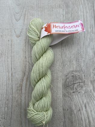 Ein handgefärbter Strang Herzfasern No. 94 – Cashmere Cloud in der Farbe Pistazie. Zartes pastelliges Grün mit leichtem Seidenglanz, fotografiert auf hellem Holz mit Herzfasern-Banderole.