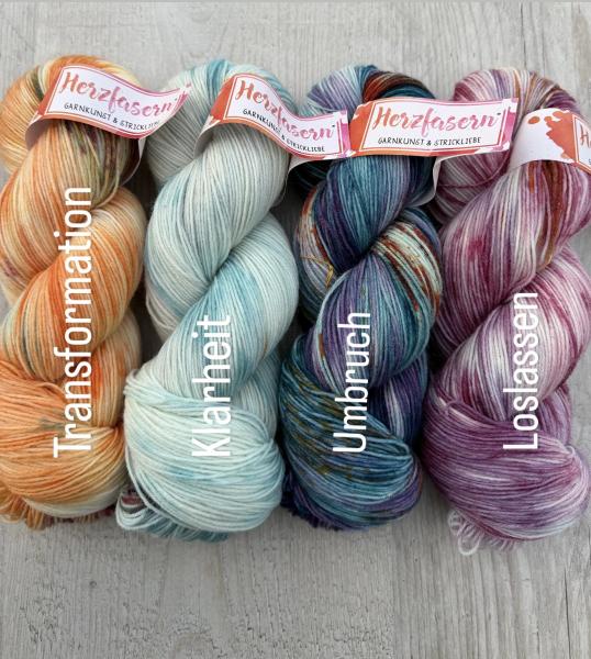 Vier handgefärbte Garnstränge der Marke Herzfasern liegen nebeneinander. Jeder Strang zeigt eine eigene Farbwelt. Zusammen wirken die Farben harmonisch und ausgewogen und bilden ein stimmiges Set.