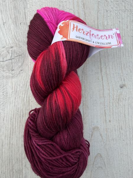 Ein handgefärbter Garnstrang in dunklen Beerentönen und Rot mit leuchtenden Pinkakzenten liegt auf einem hellen Holzuntergrund. Die Farben wirken tief und harmonisch mit lebendigen Highlights.