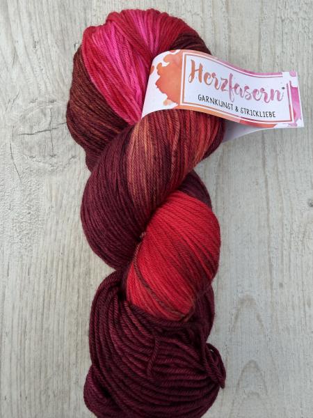 Ein handgefärbter Garnstrang in tiefen Rot- und Beerentönen mit leuchtendem Pink sowie warmen rost- und braunfarbenen Akzenten liegt auf einem hellen Holzuntergrund. Die Farben wirken lebendig und einzigartig.