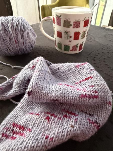 Angestricktes Strickstück aus lilafarbener Wolle mit rosafarbenen Sprenkeln auf einem Tisch, daneben ein Wollknäuel und eine Tasse Kaffee. Die Maschenstruktur ist glatt und gleichmäßig.