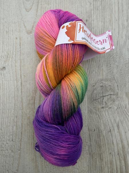 Handgefärbter Wollstrang mit lebendigen Farbverläufen in Pink, Orange, Grün und Violett auf hellem Holzhintergrund. Der Strang trägt eine Herzfasern-Banderole.