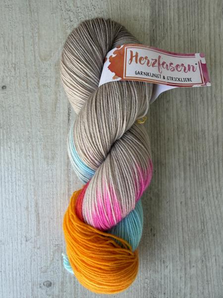 Ein handgefärbter Garnstrang in warmem Sandbeige mit Farbakzenten in Pink, Türkis und Orange liegt auf einem hellen Holzuntergrund. Der Strang ist mit einer Herzfasern-Banderole versehen.