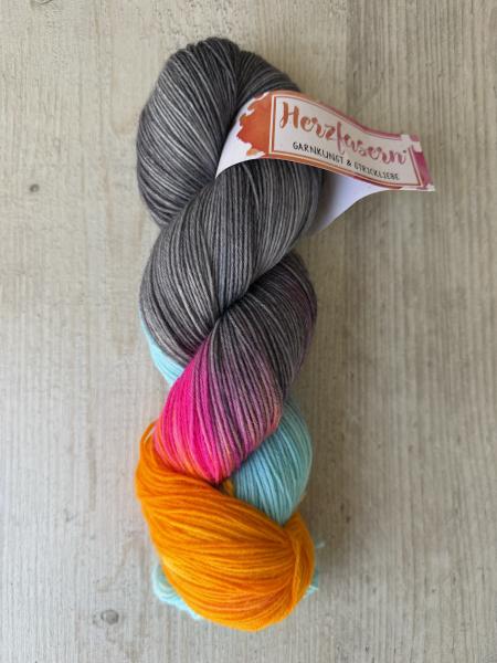 Ein handgefärbter Garnstrang in rauchigem Grau mit Farbakzenten in Pink, Türkis und Orange liegt auf einem hellen Holzuntergrund. Der Strang ist mit einer Herzfasern-Banderole versehen.