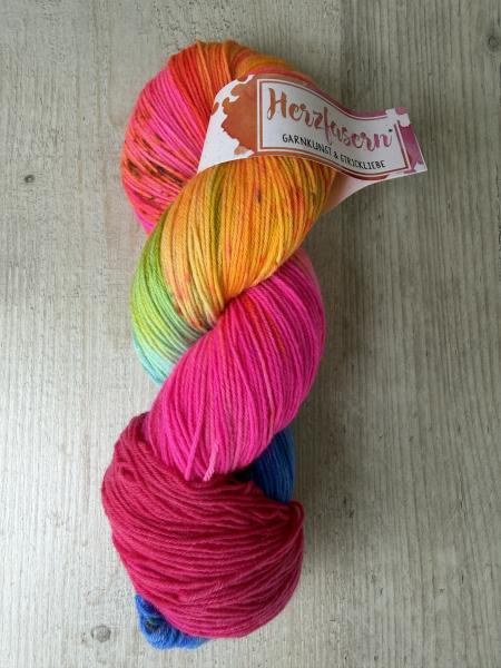 Ein handgefärbter Strang Sockenwolle von Herzfasern in kräftigen Neonfarben. Die Färbung kombiniert leuchtendes Pink, Orange, Gelb, Grün und Blau mit dunklen Sprenkeln. Der Strang liegt auf hellem Holzuntergrund und trägt eine Herzfasern-Banderole.
