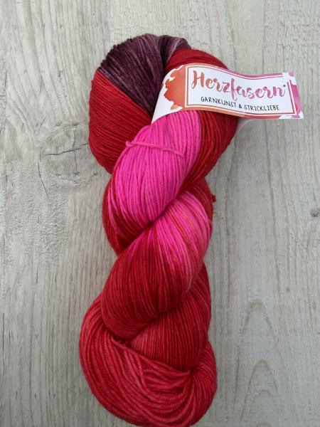 Ein handgefärbter Garnstrang in kräftigem Rot mit Übergängen zu leuchtendem Pink und Rosétönen liegt auf einem hellen Holzuntergrund. Die Farbverläufe sind weich und lebendig.