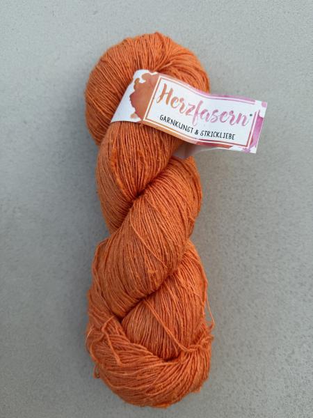 Ein handgefärbter Garnstrang aus Tussahseide in kräftigem Orange liegt auf hellem Untergrund. Die typische, leicht unregelmäßige Struktur mit kleinen Noppen ist sichtbar. Eine Herzfasern-Banderole ist am Strang befestigt.